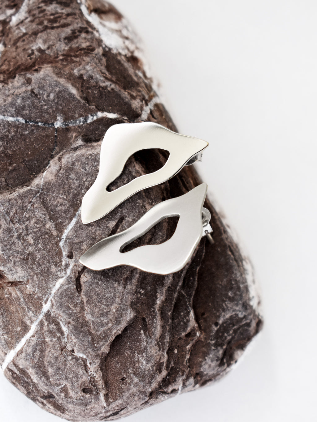 Pair of organic-shaped silver stud earrings with cutout centers on a textured stone