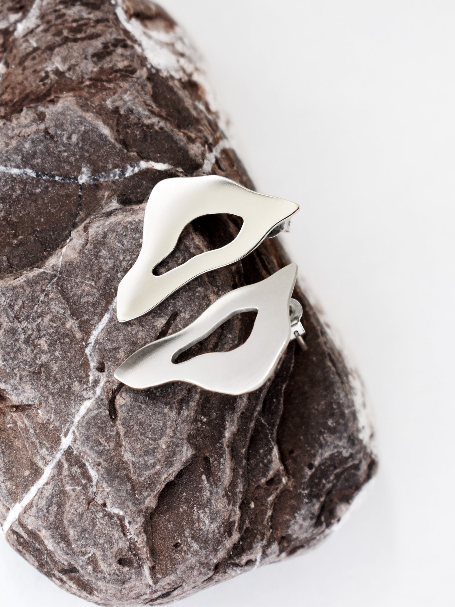 Pair of organic-shaped silver stud earrings with cutout centers on a textured stone
