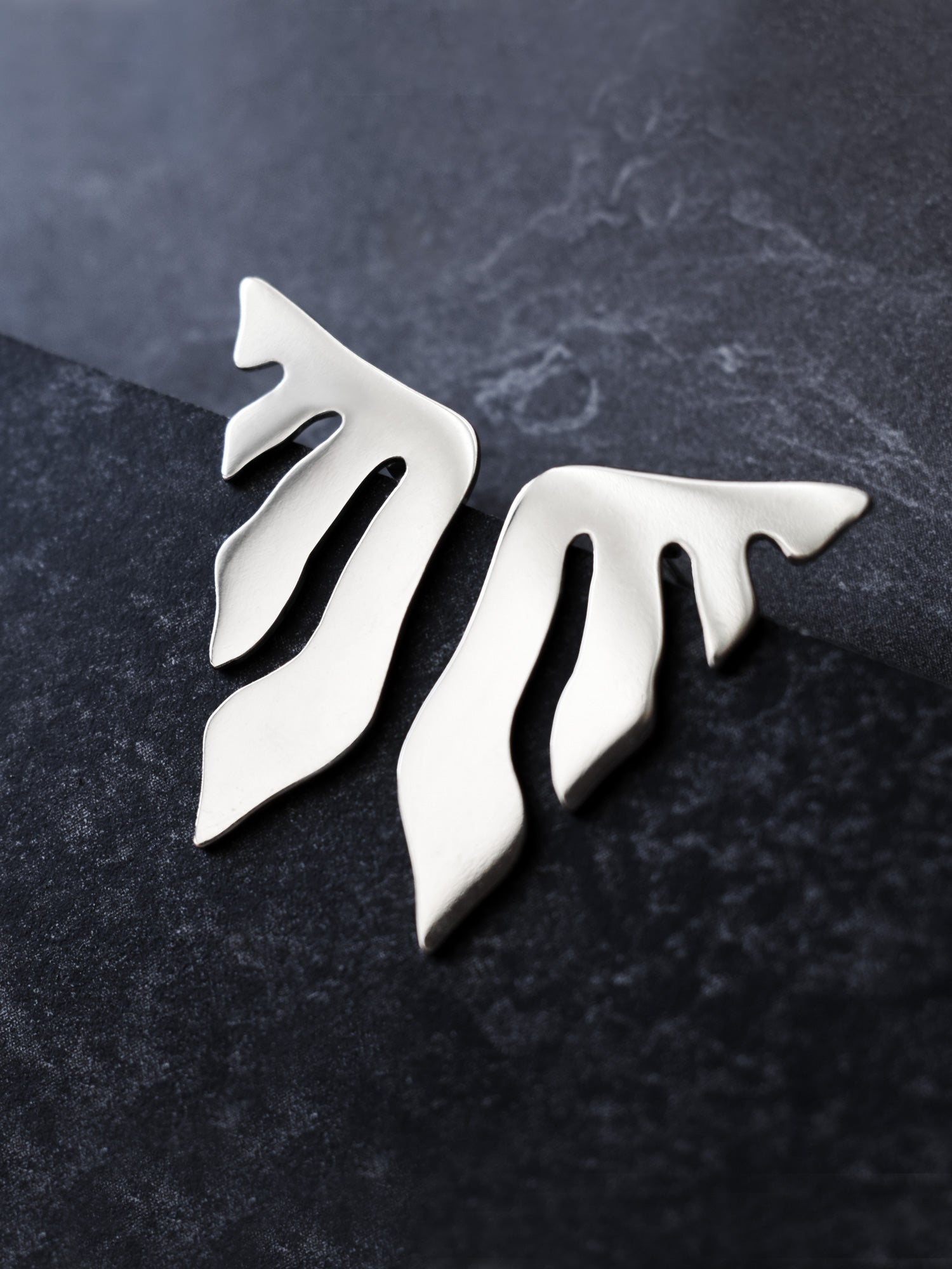 Pair of organic-shaped silver stud earrings with flowing cutouts on a dark textured surface