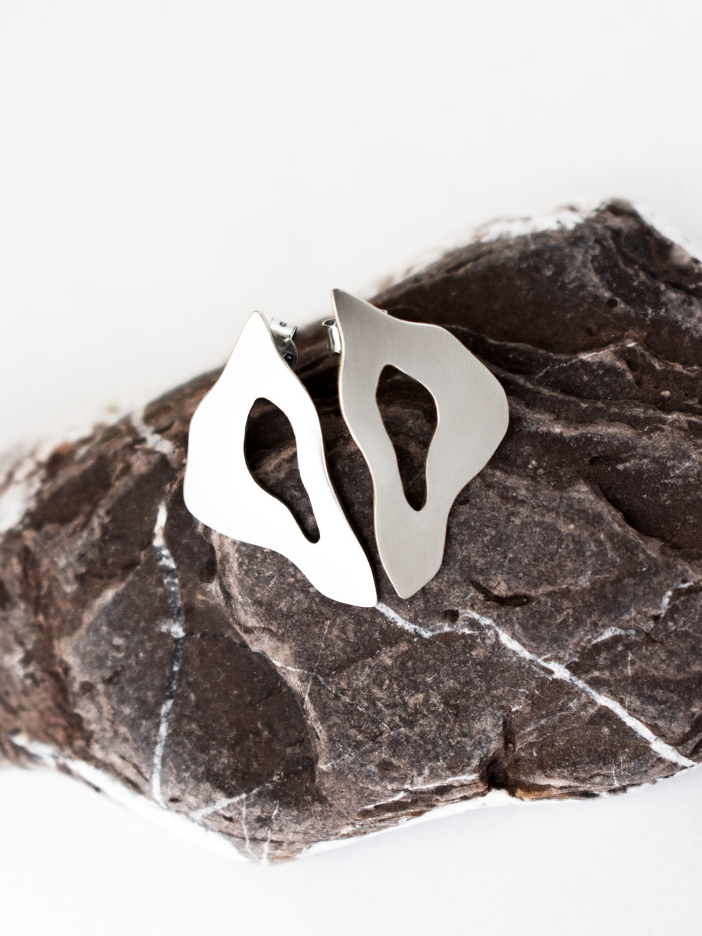 Pair of organic-shaped silver stud earrings with cutout centers on a textured stone