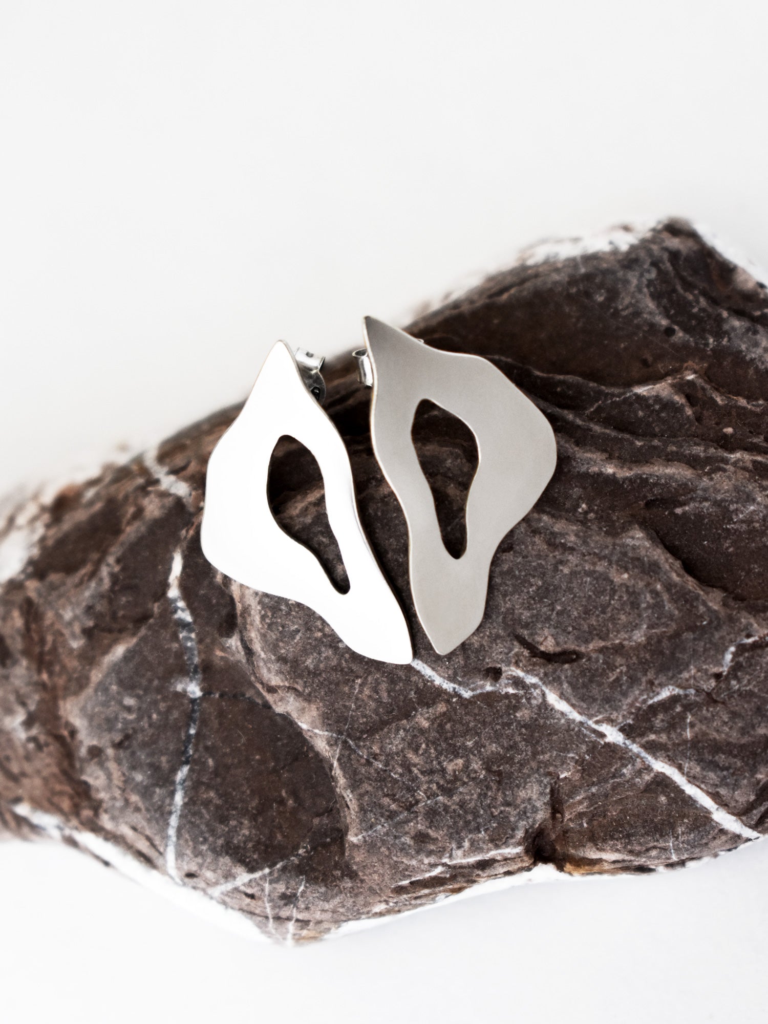 Pair of organic-shaped silver stud earrings with cutout centers on a textured stone