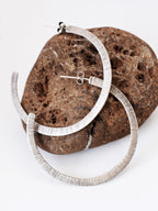 Pair of textured silver hoop earrings on a brown stone against a white background