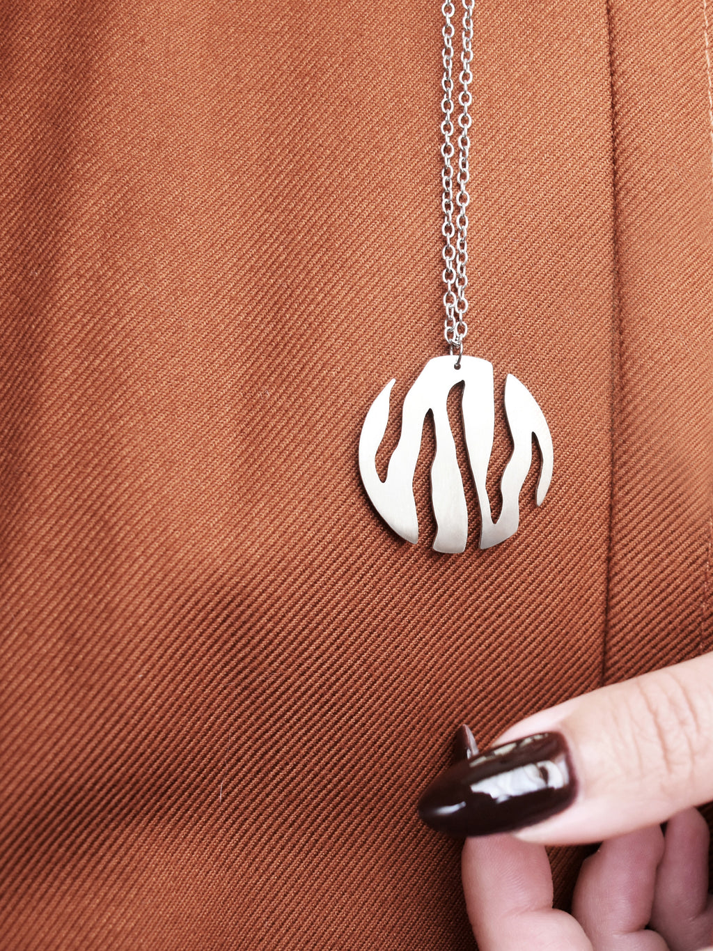 Round silver pendant with flowing cutouts and chain worn on a rust-colored garment