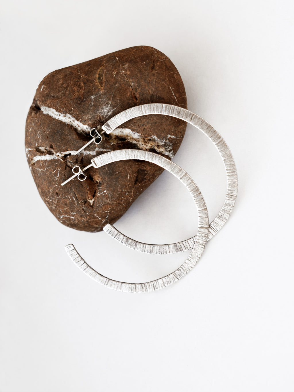 Pair of textured silver hoop earrings on a brown stone against a white background