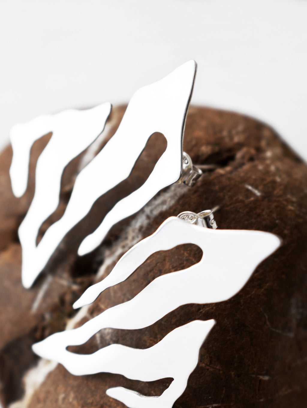 Close-up of pair of abstract-shaped silver stud earrings with flowing cutouts on a textured brown stone