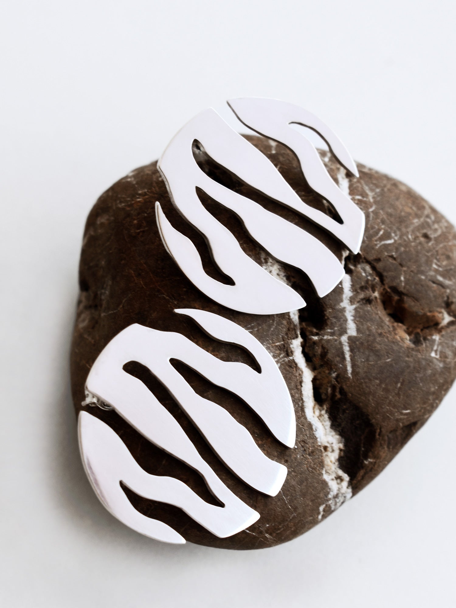 Pair of round silver stud earrings with flowing cutouts on a textured brown stone