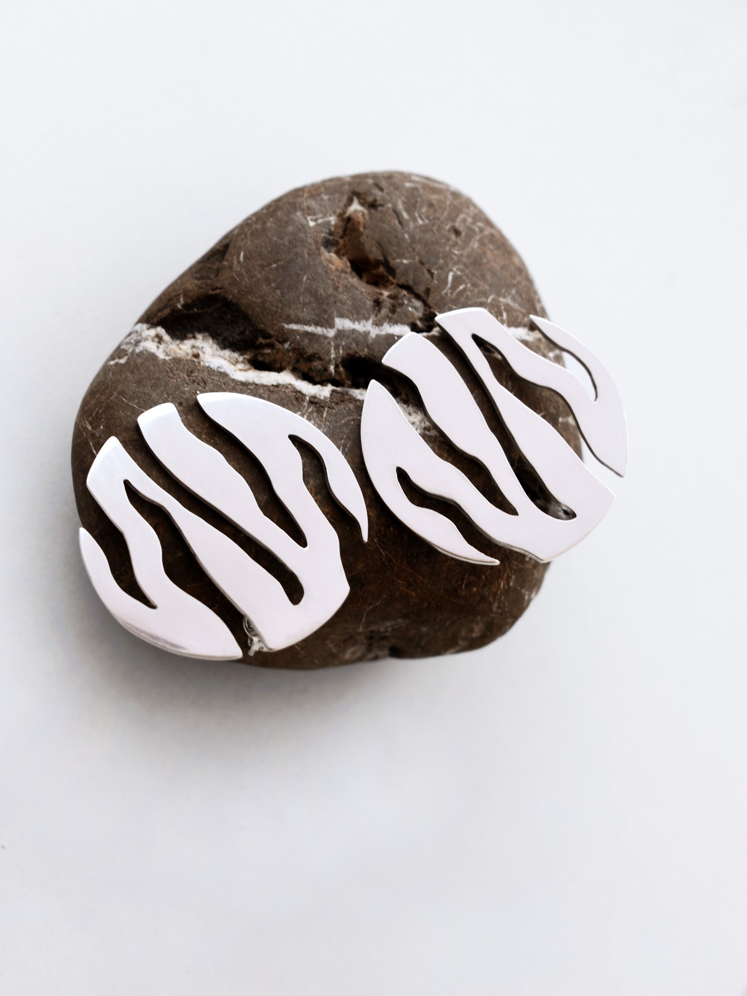 Pair of round silver stud earrings with flowing cutouts on a textured brown stone