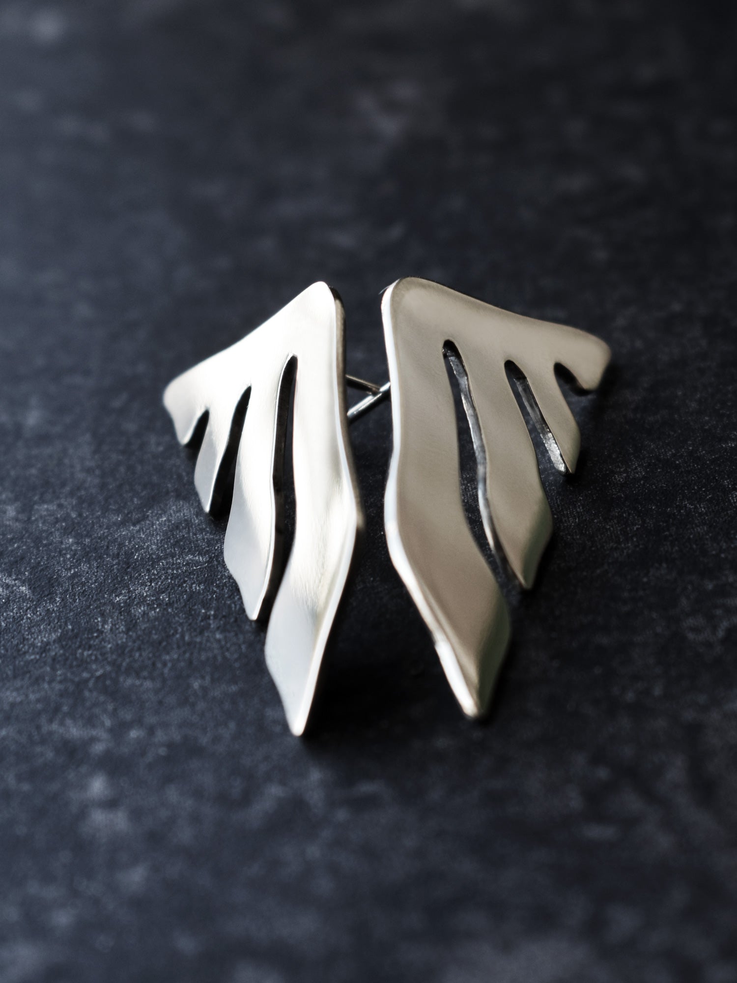 Pair of organic-shaped silver stud earrings with flowing cutouts on a dark textured surface
