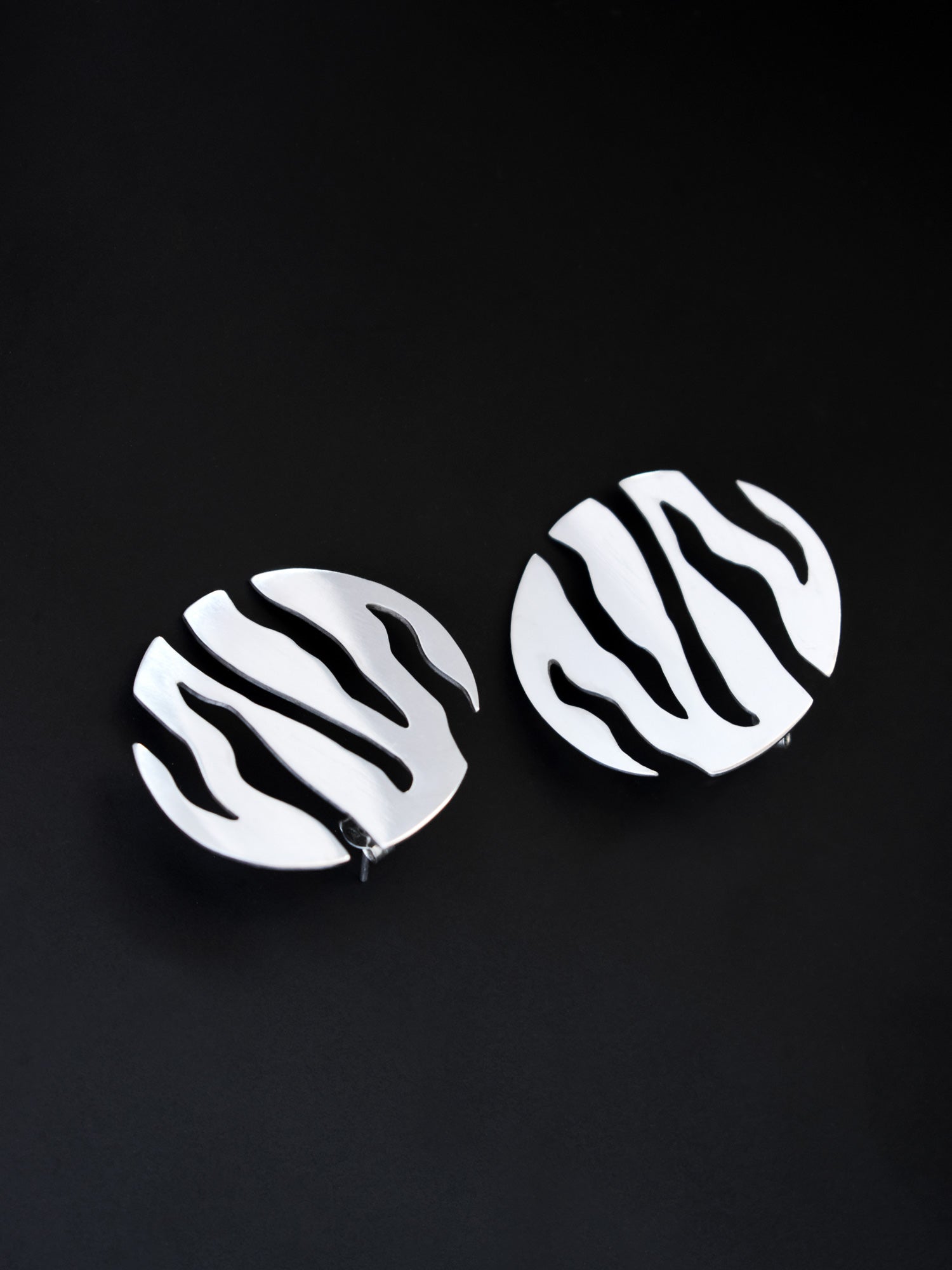 Pair of round silver stud earrings with flowing cutouts on a black background