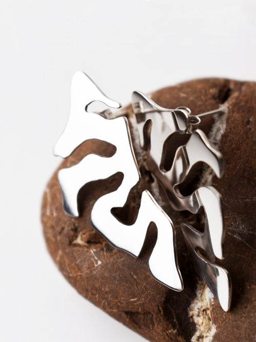 Close-up of pair of organic-shaped silver stud earrings with cutout details on a textured brown stone