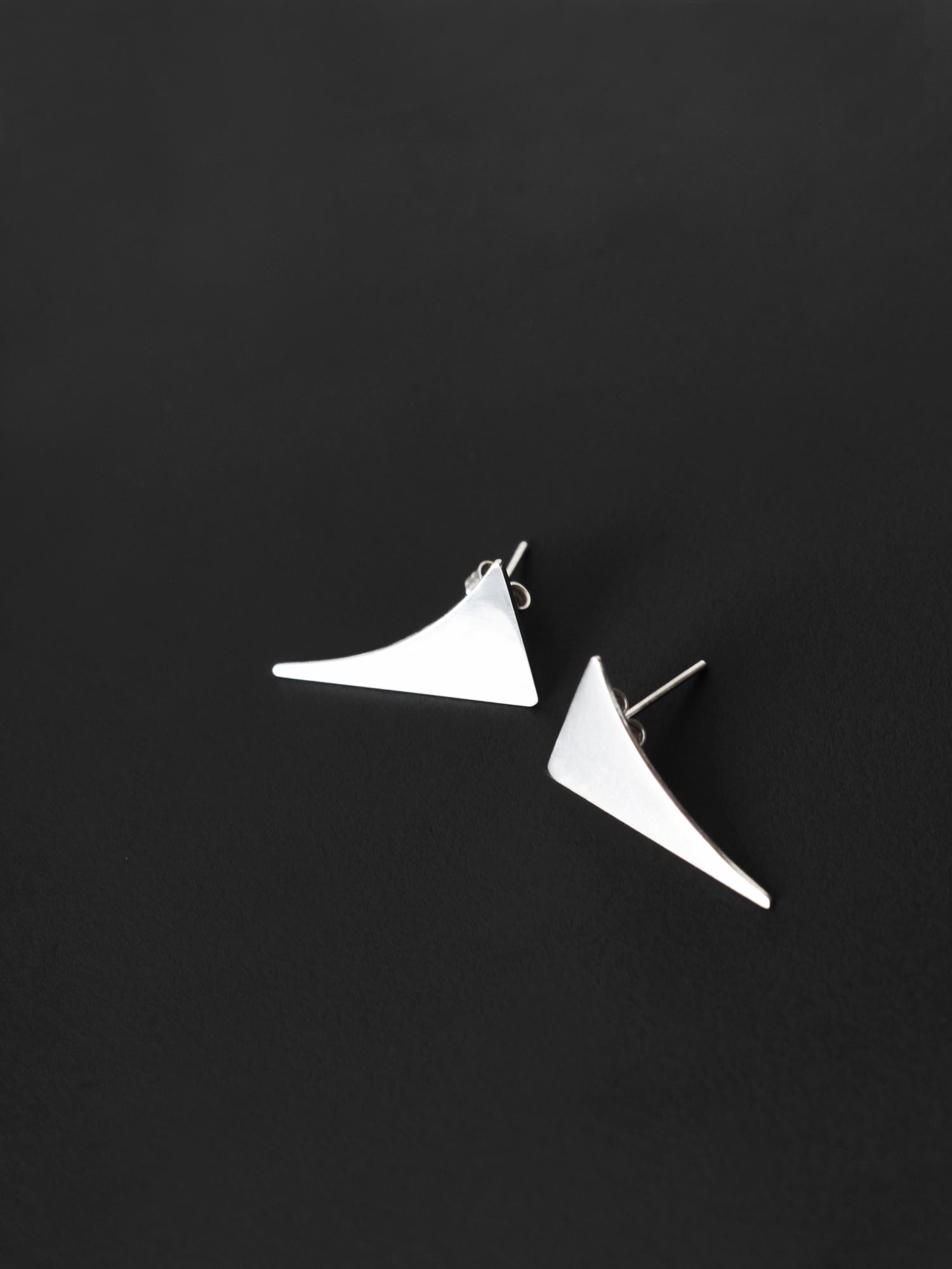 Pair of small curved triangular silver stud earrings on a black textured background