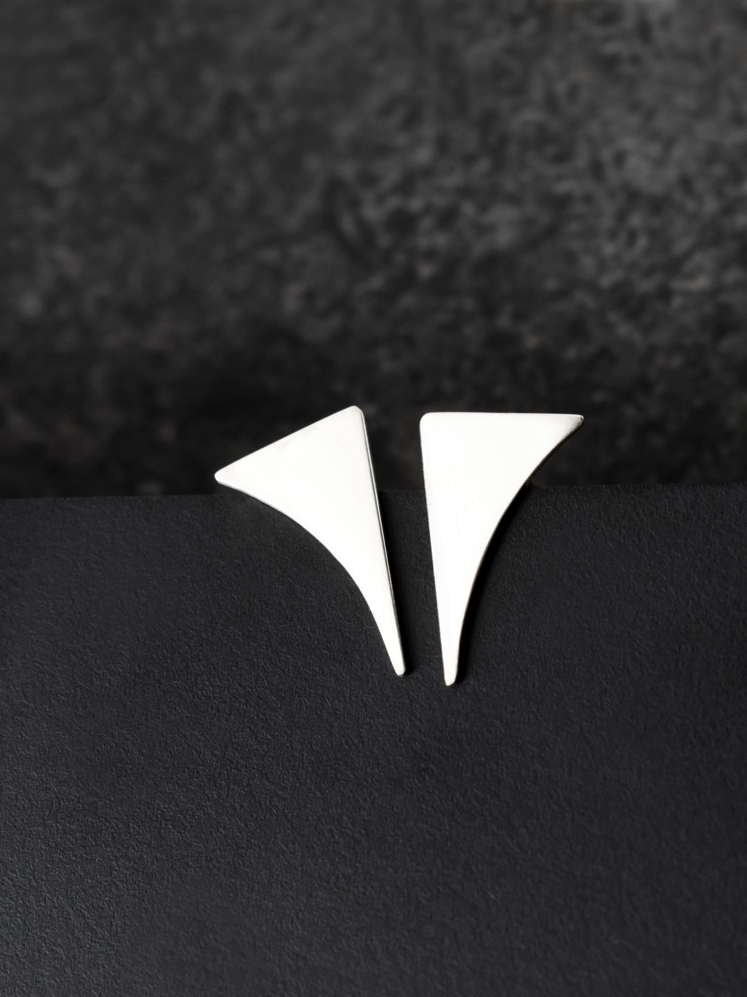 Pair of small curved triangular silver stud earrings on a black textured background